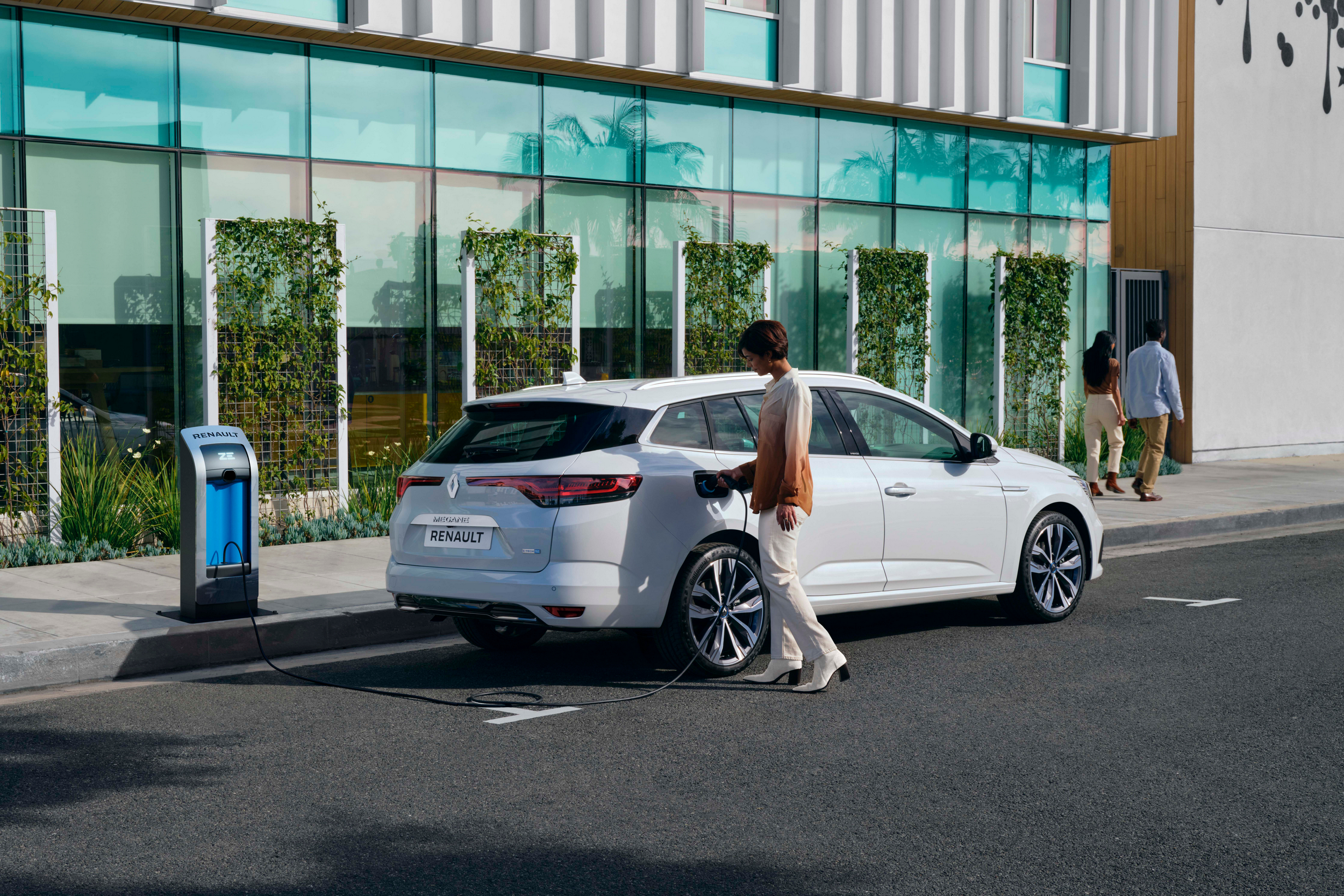 Moteris prijungia baltą įkraunamą hibridinį Renault Megane GrandTour E-Tech Plug-in Hybrid prie įkrovimo stotelės mieste