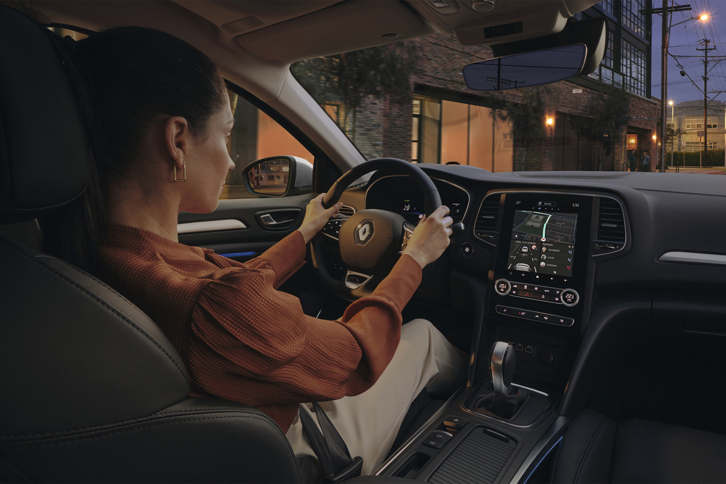 Moteris vairuoja hečbeką Renault MEGANE mieste, matomas automobilio interjeras: vairas, prietaisų skydas, centrinis ekranas ir centrinė konsolė